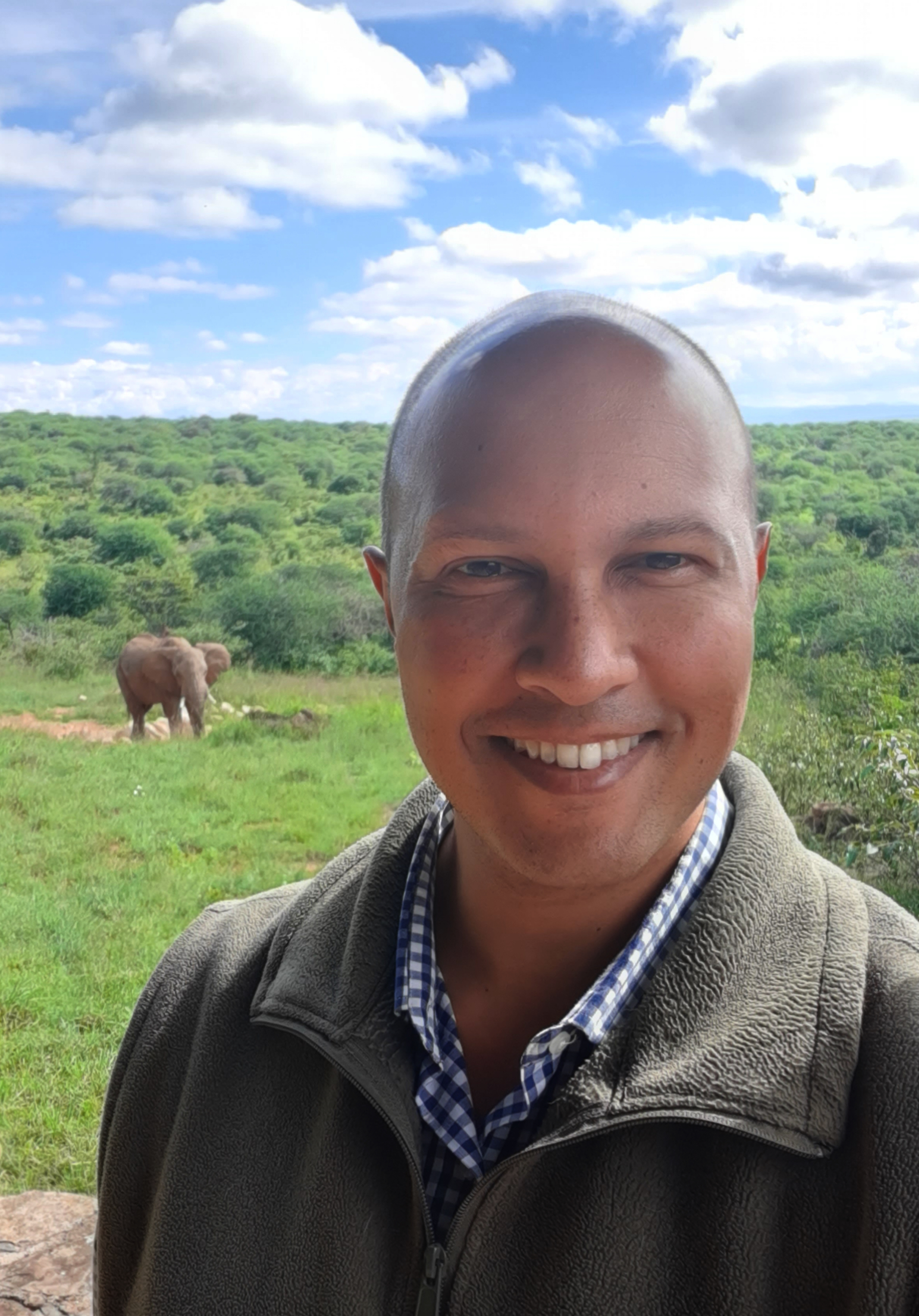 Dr. Dino Martins in the field at Mpala, Kenya, with an elephant grazing in the background